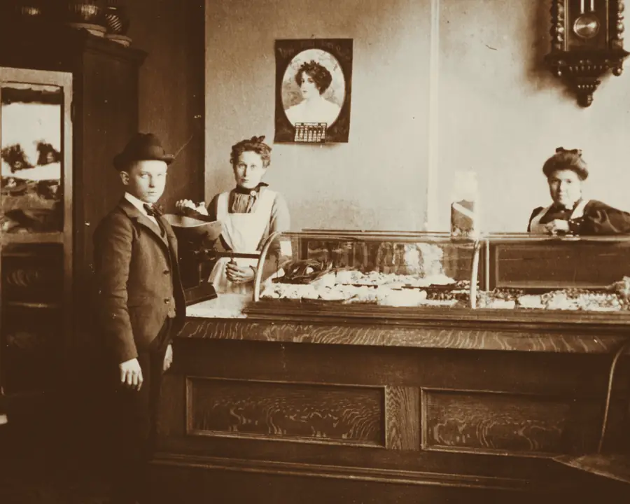 Archival photograph of another bakery: three women and a boy at a long wooden counter, behind a glass case of baked goods.