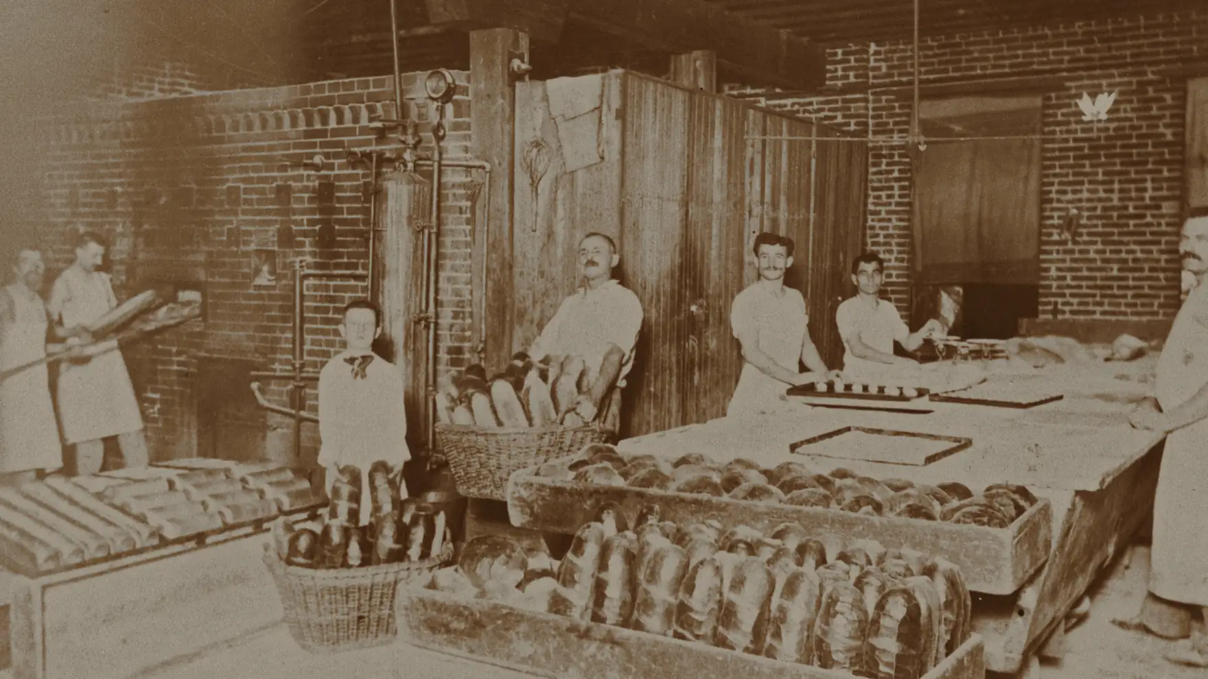 Archival photograph of another bakery: six men and a boy working a brick-oven bakehouse, drawing loaves out on a peel and filling baskets and trays.